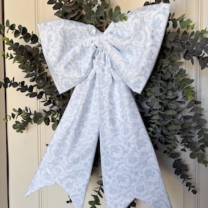 May include: A large, decorative bow made of white fabric with a light blue floral pattern. The bow is centered on a wreath of green eucalyptus leaves, creating a classic and elegant look. The bow's tails have pointed ends.
