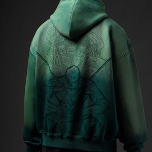 Puede incluir: Sudadera con capucha verde con efecto degradado, con una ilustraci&oacute;n detallada en la espalda. La obra de arte representa una figura con espadas y un grupo de caras. La sudadera tiene capucha y un corte holgado.