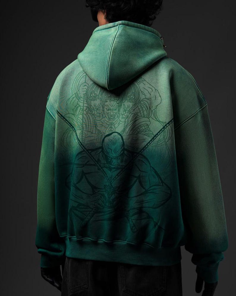 Puede incluir: Sudadera con capucha verde con efecto degradado, con una ilustraci&oacute;n detallada en la espalda. La obra de arte representa una figura con espadas y un grupo de caras. La sudadera tiene capucha y un corte holgado.