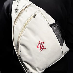 Puede incluir: Bandolera de color crema con cremallera diagonal y bolsillo lateral de malla. La bolsa presenta un diseño bordado en rojo y un charm plateado. La bolsa se lleva sobre el hombro.