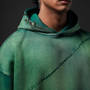 Puede incluir: Sudadera con capucha verde con un dise&ntilde;o de costura &uacute;nico en la parte delantera. La sudadera con capucha tiene una capucha con cord&oacute;n y anillos met&aacute;licos dorados. La tela tiene un aspecto descolorido y texturizado, con tonos verdes m&aacute;s oscuros en los bordes.