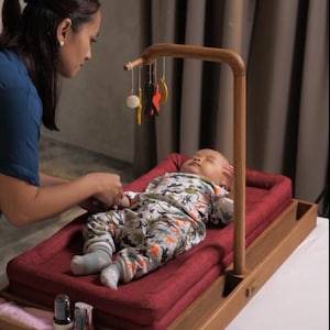 May include: A wooden baby changing station with a red cushion and a mobile. The baby is wearing a dinosaur-patterned onesie. The station has a lower shelf with a pink towel and bottles.