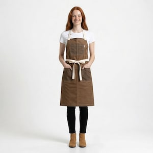 May include: A brown apron with a cream-colored neck strap and waist tie. The apron has two large front pockets and a small pocket on the chest. The model is wearing a white t-shirt, black pants, and brown boots.