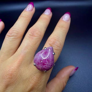 May include: A teardrop-shaped, deep magenta gemstone ring set in a silver-toned metal prong setting. The stone features a textured, natural surface with visible crystalline inclusions and a polished finish, secured by four small metal prongs.