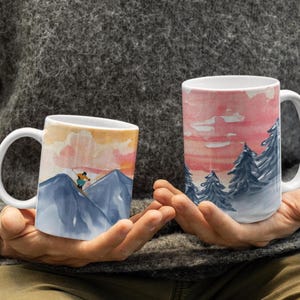 May include: Two white ceramic mugs with wraparound watercolor designs. One mug depicts a skier on a mountain with a sunset sky. The other mug shows a forest of blue trees against a pink and red sky. Both mugs have white handles.