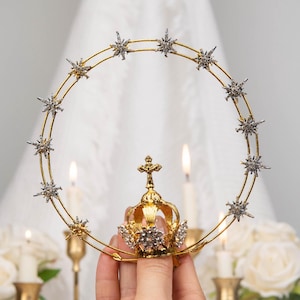 Op de afbeelding: Een goudkleurig, kroonvormig decoratief stuk met een cirkelvormig ontwerp, versierd met stervormige versieringen. De kroon heeft een kleinere, sierlijke kroon aan de basis en wordt gehouden voor een witte achtergrond met kaarsen en bloemen.
