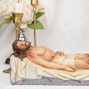 Può includere: Una statuetta in resina dipinta a mano raffigurante una figura distesa con lunghi capelli castani e barba, adagiata su un panno bianco drappeggiato sopra una base grigia con texture in pietra. La figura presenta segni rossi visibili sulla pelle.