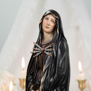 May include: A religious figurine depicting a figure in a black hooded robe with gold trim, featuring a heart pierced by seven silver swords on the chest. The figure has a sorrowful expression with visible tears and hands clasped together.