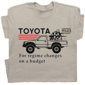 May include: Beige t-shirt featuring a black graphic of a Toyota Hilux truck with a mounted weapon. The text "TOYOTA" and "HILUX" are above the truck, with the phrase "For regime changes on a budget" below.