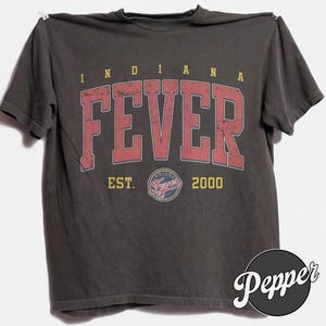 May include: A charcoal gray short-sleeve t-shirt featuring the text INDIANA FEVER in large, distressed red block letters. Below the text, the shirt displays the words EST. 2000 flanking a circular Indiana Fever team logo in red, blue, and white.