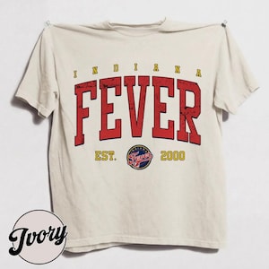 May include: An ivory short-sleeve t-shirt featuring the text INDIANA FEVER in large, distressed red block letters. Below the text is the circular Indiana Fever team logo, flanked by the yellow text EST. and 2000 on either side of the graphic.