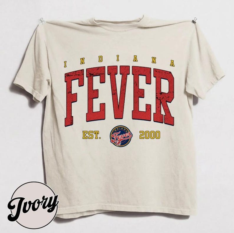 May include: An ivory short-sleeve t-shirt featuring the text INDIANA FEVER in large, distressed red block letters. Below the text is the circular Indiana Fever team logo, flanked by the yellow text EST. and 2000 on either side of the graphic.
