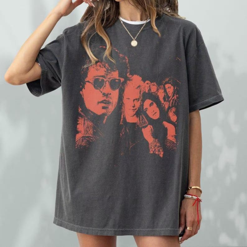 May include: A charcoal gray short-sleeve t-shirt featuring a faded red graphic print of a group of people, including one person wearing sunglasses. The shirt has a crew neckline and a loose, relaxed fit, showcasing a vintage-inspired aesthetic.