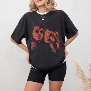 May include: A black short-sleeve t-shirt featuring a prominent red-toned graphic print of a group of people, including a central figure wearing sunglasses. The shirt has a crew neckline and a loose, relaxed fit, showcasing a detailed illustration.
