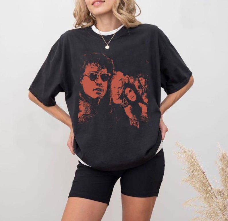 May include: A black short-sleeve t-shirt featuring a prominent red-toned graphic print of a group of people, including a central figure wearing sunglasses. The shirt has a crew neckline and a loose, relaxed fit, showcasing a detailed illustration.