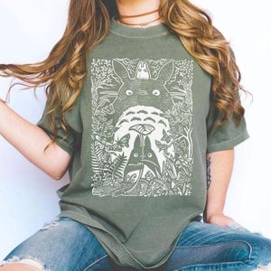 Puede incluir: Camiseta verde salvia con un gr&aacute;fico blanco de los personajes de la pel&iacute;cula de animaci&oacute;n, Mi vecino Totoro. El dise&ntilde;o incluye el Totoro grande, Totoros m&aacute;s peque&ntilde;os, mariposas y follaje. La camiseta es de cuello redondo.