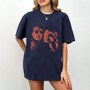 May include: A dark navy blue short-sleeve t-shirt featuring a graphic print in shades of red and orange. The design displays a group of people, including a prominent figure wearing sunglasses, arranged in a layered, artistic composition.