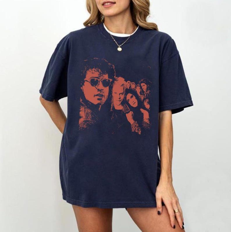 May include: A dark navy blue short-sleeve t-shirt featuring a graphic print in shades of red and orange. The design displays a group of people, including a prominent figure wearing sunglasses, arranged in a layered, artistic composition.