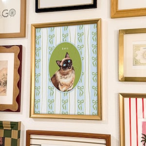 Puede incluir: Una impresión enmarcada con un retrato de gato siamés dentro de un óvalo verde, sobre un fondo a rayas azules y blancas con lazos. La impresión está en un marco dorado, expuesta entre otras obras de arte enmarcadas.