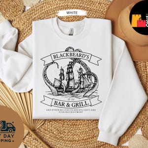 May include: White sweatshirt with a black and white graphic of a pirate ship being attacked by a giant squid. The text on the sweatshirt reads "BLACKBEARD'S BAR & GRILL" and "EST 1717". The sweatshirt also has the text "AND OTHER DELICACIES AND DELIGHTS AND FISHING EQUIPMENT".