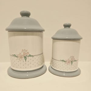 May include: Two ceramic canisters with pale blue lids and bases. The white bodies feature a floral design with pink flowers and green leaves. A patterned band with small crosses accents the base of each canister.