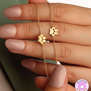 May include: A delicate gold-colored chain bracelet featuring two paw-shaped charms. The charms have a cutout design and the word "Teddy" is engraved on one. The bracelet is held between fingers with a neutral-toned manicure.