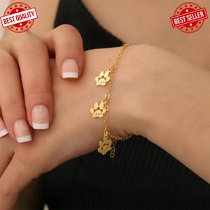 Puede incluir: Pulsera dorada con tres colgantes de huellas de pata. Cada colgante tiene un nombre grabado: "Lola" y "Miller". La pulsera se lleva en la muñeca. La imagen también incluye un sello de "Best Quality" y "Best Seller".