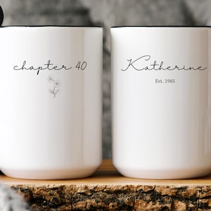 May include: Two white ceramic mugs with black handles and rims. One mug reads "chapter 40" with a floral illustration, the other says "Katherine Est. 1985" in script. The mugs are on a wooden surface.