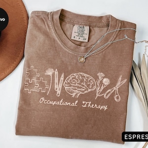 Pu&ograve; includere: T-shirt color espresso con un design grafico bianco raffigurante pezzi di puzzle, matite, un cervello, fiori e forbici. Le parole "Occupational Therapy" sono sotto la grafica. Una collana d'argento con un ciondolo &egrave; sulla maglietta.