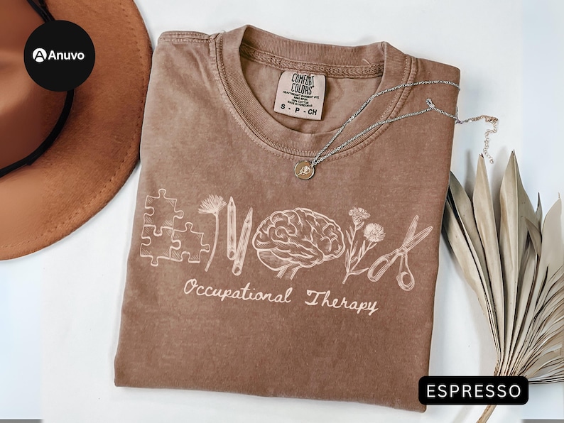 Pu&ograve; includere: T-shirt color espresso con un design grafico bianco raffigurante pezzi di puzzle, matite, un cervello, fiori e forbici. Le parole "Occupational Therapy" sono sotto la grafica. Una collana d'argento con un ciondolo &egrave; sulla maglietta.