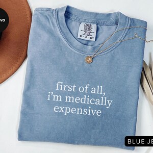 May include: A blue jean colored t-shirt with the text "first of all, I'm medically expensive" in white. The shirt is made by Comfort Colors and is size S-P-CH. A gold necklace with a pendant is on the shirt.
