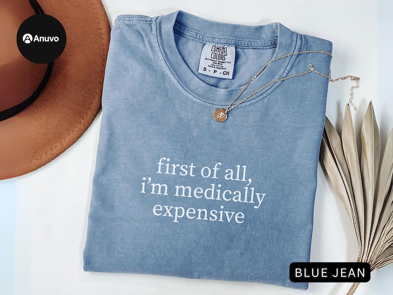 May include: A blue jean colored t-shirt with the text "first of all, I'm medically expensive" in white. The shirt is made by Comfort Colors and is size S-P-CH. A gold necklace with a pendant is on the shirt.