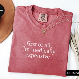 May include: Crimson-colored t-shirt with the text "first of all, i'm medically expensive" in white. A gold pendant necklace rests on the shirt. The shirt is folded on a white surface, with a brown hat and dried plants in the background.
