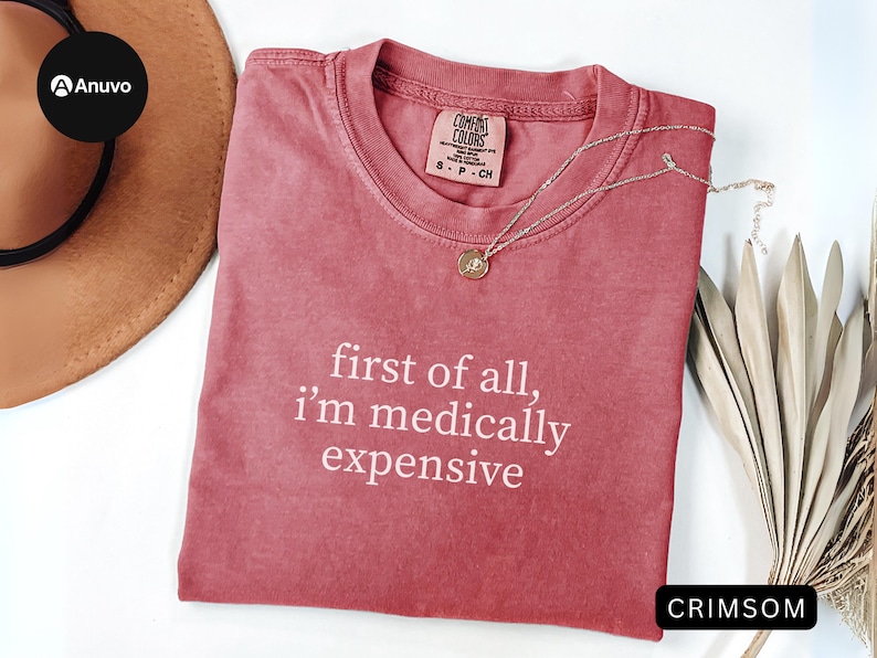 May include: Crimson-colored t-shirt with the text "first of all, i'm medically expensive" in white. A gold pendant necklace rests on the shirt. The shirt is folded on a white surface, with a brown hat and dried plants in the background.