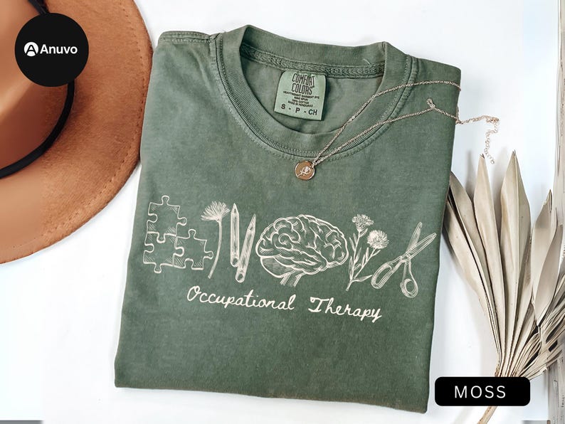 May include: A moss green t-shirt with white graphic design. The design includes puzzle pieces, pencils, a brain, flowers, and scissors, with the words 'Occupational Therapy' below. A gold necklace with a pendant is on the shirt. The shirt is on a white surface with a brown hat and dried leaves.