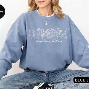 Pu&ograve; includere: Una felpa azzurra con le parole "Occupational Therapy" e grafica associata in bianco. Il design include un pezzo di puzzle, un fiore, un cervello e delle forbici. La persona indossa jeans blu.