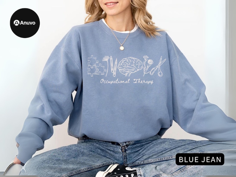 Pu&ograve; includere: Una felpa azzurra con le parole "Occupational Therapy" e grafica associata in bianco. Il design include un pezzo di puzzle, un fiore, un cervello e delle forbici. La persona indossa jeans blu.