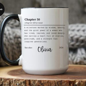 May include: White ceramic mug with a black handle and rim. The mug features the text "Chapter 50" with a definition, the name "Olivia", and the year "2026". The mug is on a wooden surface.