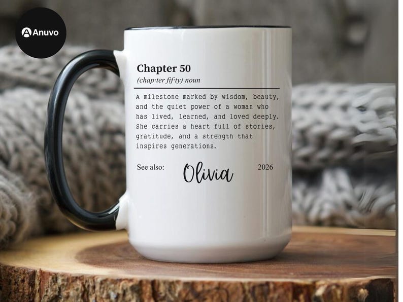 May include: White ceramic mug with a black handle and rim. The mug features the text "Chapter 50" with a definition, the name "Olivia", and the year "2026". The mug is on a wooden surface.
