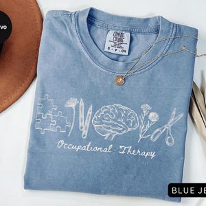 Pu&ograve; includere: Una maglietta blu jeans Comfort Colors con elementi grafici bianchi relativi alla terapia occupazionale. Il design include pezzi di puzzle, matite, un cervello, fiori e forbici. Una collana d'oro con un ciondolo &egrave; appoggiata sulla maglietta.