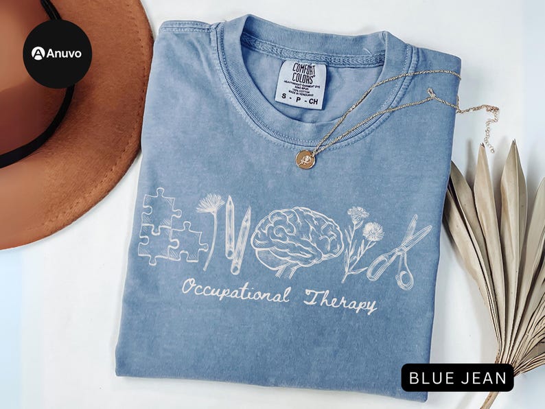 Pu&ograve; includere: Una maglietta blu jeans Comfort Colors con elementi grafici bianchi relativi alla terapia occupazionale. Il design include pezzi di puzzle, matite, un cervello, fiori e forbici. Una collana d'oro con un ciondolo &egrave; appoggiata sulla maglietta.