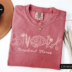 Pu&ograve; includere: T-shirt color cremisi con le parole "Occupational Therapy" e grafica correlata. Il design include un puzzle, matite, un cervello, fiori e forbici. Una collana d'argento con un ciondolo poggia sulla maglietta.
