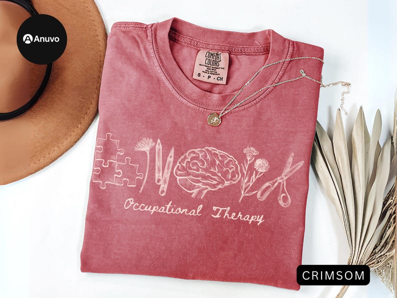 Pu&ograve; includere: T-shirt color cremisi con le parole "Occupational Therapy" e grafica correlata. Il design include un puzzle, matite, un cervello, fiori e forbici. Una collana d'argento con un ciondolo poggia sulla maglietta.