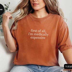 May include: Rust-colored t-shirt with the text "first of all, I'm medically expensive" in white. The shirt has short sleeves and a relaxed fit. The model is wearing blue jeans.