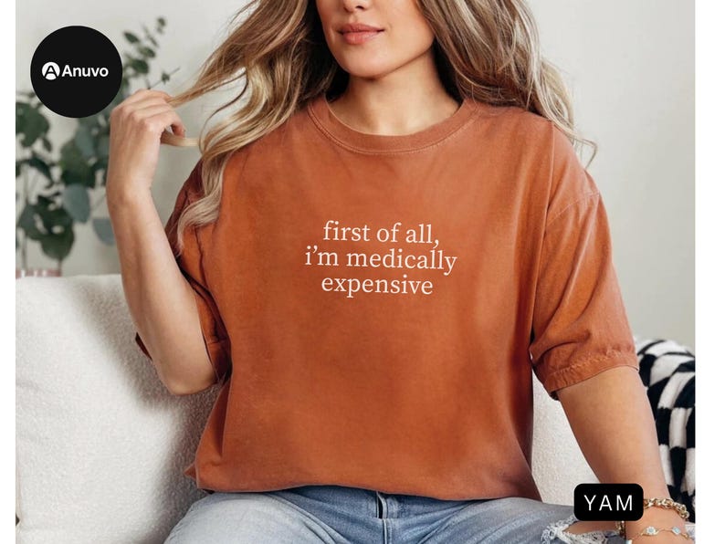 May include: Rust-colored t-shirt with the text "first of all, I'm medically expensive" in white. The shirt has short sleeves and a relaxed fit. The model is wearing blue jeans.