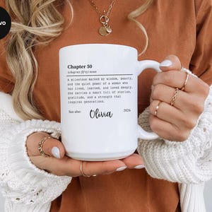 May include: White ceramic mug with the text "Chapter 50" and a definition. The mug also includes the name "Olivia" and the year "2026". The mug is held by a person wearing a white sweater and gold rings.