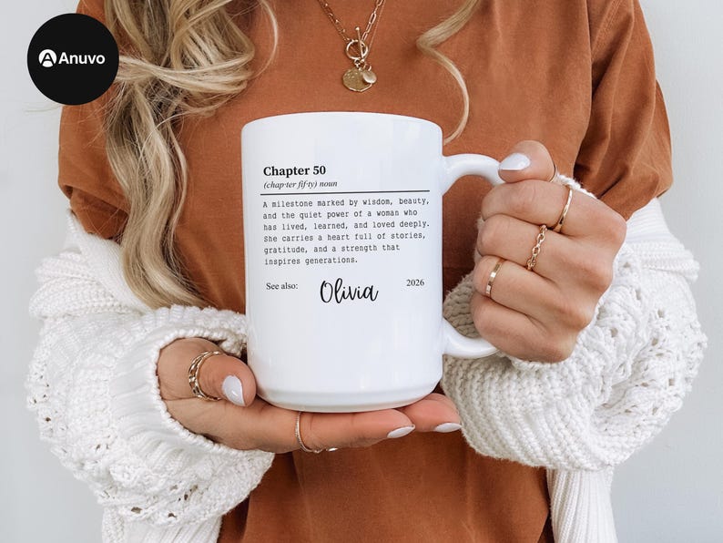 May include: White ceramic mug with the text "Chapter 50" and a definition. The mug also includes the name "Olivia" and the year "2026". The mug is held by a person wearing a white sweater and gold rings.