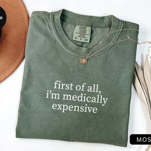 May include: Moss green t-shirt with the text "first of all, I'm medically expensive" in white. A gold necklace with a pendant rests on the shirt. The shirt is folded and displayed with a brown hat and dried plant stems.