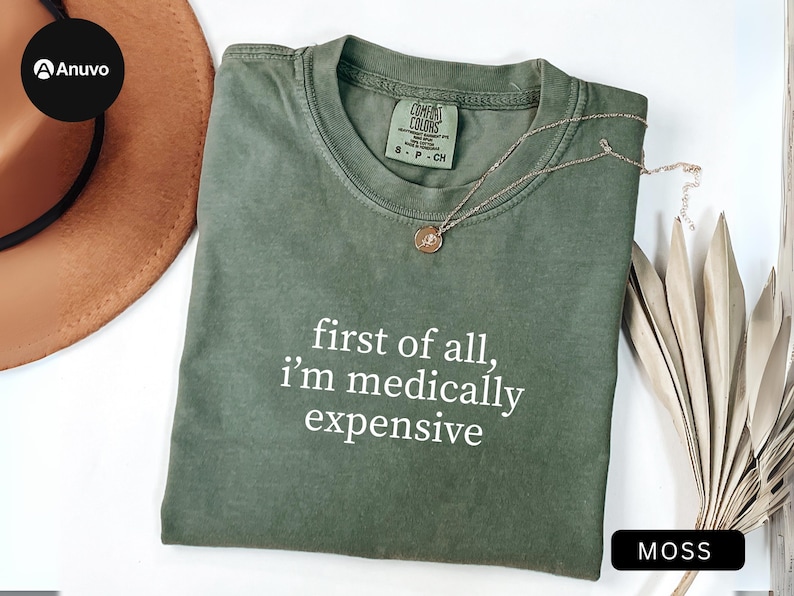 May include: Moss green t-shirt with the text "first of all, I'm medically expensive" in white. A gold necklace with a pendant rests on the shirt. The shirt is folded and displayed with a brown hat and dried plant stems.