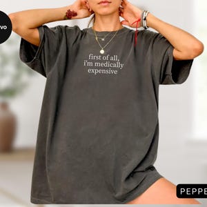 May include: A dark gray oversized t-shirt with the text "first of all, i'm medically expensive" in white. The shirt has short sleeves and a relaxed fit. The word "PEPPER" is in a black rectangle in the bottom right corner.